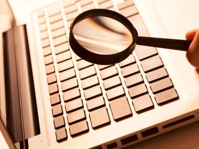 Hand holding a magnifying glass over a laptop keyboard, stock research tools article
